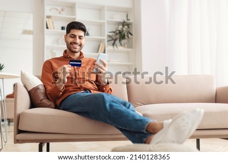 Internet Mobile Shopping Concept. Positive Middle Eastern male using cell phone holding looking at credit card purchasing things online sitting on sofa buying gifts in web store, full body length Royalty-Free Stock Photo #2140128365