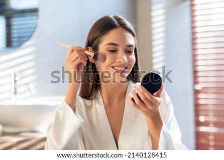 Daily Makeup. Smiling Young Female Applying Blush With Makeup Brush In Bathroom, Beautiful Happy Woman Wearing White Silk Robe Using New Cosmetics While Getting Ready At Home, Copy Space Royalty-Free Stock Photo #2140128415