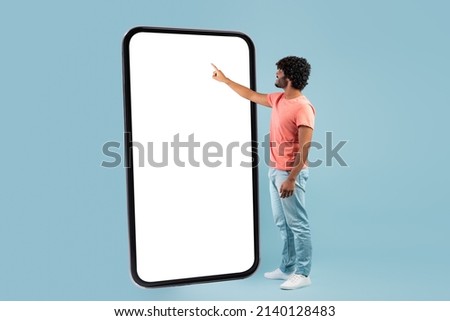 Stylish curly young hindu guy in casual outift touching huge smartphone white empty screen over blue studio background, checking newest mobile app, panorama with copy space, mockup Royalty-Free Stock Photo #2140128483