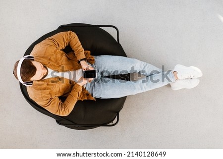 Man Using Smartphone Texting And Browsing Internet Wearing Headphones, Listening To Music Online Or Podcast Sitting In Chair Indoors. Above View Shot. Gadgets And Technology Concept