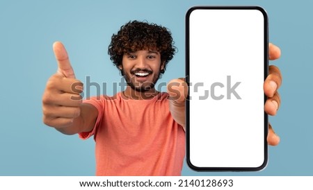 Handsome indian millennial guy showing brand new smartphone with empty white screen and thumb up, smiling at camera over blue studio background, mockup, mobile application concept, panorama Royalty-Free Stock Photo #2140128693