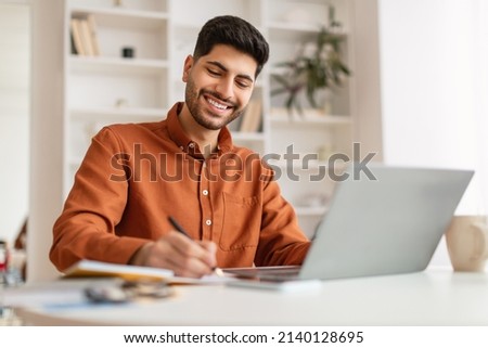 Smiling Arab Man Using Laptop Working With Documents At Home Office, Handsome Eastern Male Entrepreneur Sitting At Desk With Computer, Checking Financial Reports And Taking Notes, Free Copy Space