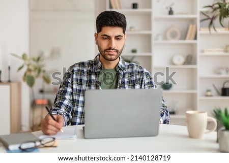 Focused Busy Arab Man Using Laptop Working With Documents At Home Office, Handsome Eastern Male Entrepreneur Sitting At Desk With Computer, Checking Financial Reports And Taking Notes, Free Copy Space