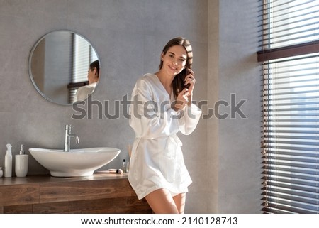 Portrait Of Beautiful Young Female Wearing White Silk Robe Posing In Luxury Bathroom Interior, Attractive Millennial Woman Making Beauty Treatments At Home, Touching Her Hair And Smiling At Camera Royalty-Free Stock Photo #2140128743