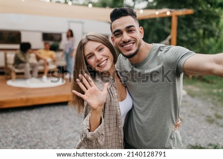 Happy loving multiracial couple taking selfie near camper van, resting with friends outdoors, free space. Arab guy and his girlfriend having fun summer camping trip, making mobile photo