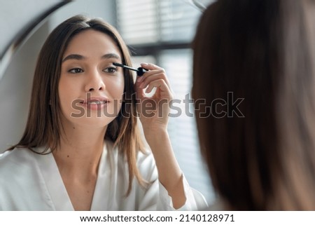 Beautiful Millennial Lady Doing Daily Makeup Near Mirror In Bathroom, Attractive Young Woman Applying Mascara On Eyelashes, Holding Brush And Smiling To Her Reflection, Selective Focus, Closeup Royalty-Free Stock Photo #2140128971