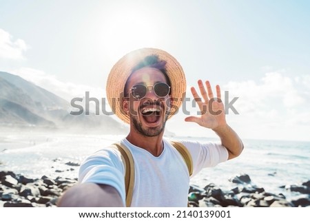 Young man with backpack taking selfie portrait outside - Smiling happy guy enjoying summer holidays at the beach - Millennial showing victory hands symbol to the camera - Youth and journey Royalty-Free Stock Photo #2140139001