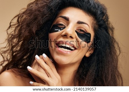 Smiling tanner pretty Latin female with black hydrogel patches under eye laugh at camera posing isolated over pastel beige background. Cosmetic product ad Natural beauty concept Studio portrait