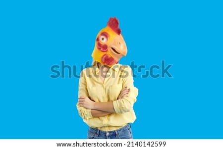 Person in yellow shirt and chicken mask on head standing with arms folded, isolated on blue studio background. Anonymous female with rubber mask of rooster feeling confusion. Creative advertising shot