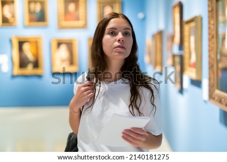 Focused young girl visitor holds a booklet with an exhibition program while admiring the paintings in the museum Royalty-Free Stock Photo #2140181275