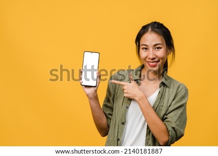 Young Asia lady show empty smartphone screen with positive expression, smiles broadly, dressed in casual clothing feeling happiness on yellow background. Mobile phone with white screen in female hand.