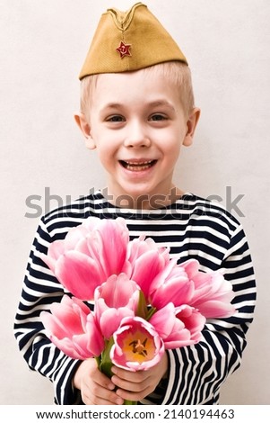 Cute smiling boy in Soviet military cap with a bouquet of pink tulips in striped t-shirt on light background. Holiday day of victory, May 9, Fatherland day. Vertical banner, photo