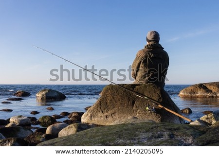 Back view of a sea trout angler with spinning rod sitting on a rock. Royalty-Free Stock Photo #2140205095