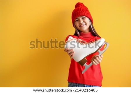 Little Asian girl in warm clothes with ice skate on yellow background
