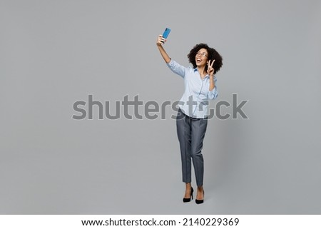 Full body young employee business corporate lawyer woman of African American ethnicity in formal shirt work in office doing selfie shot on mobile cell phone show v-sign isolated on grey background.