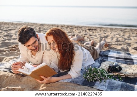 Smiling sunlit young couple two family man woman in white clothes hug lying on picnic plaid read book rest relax together at sunrise over sea beach ocean outdoor seaside in summer day sunset evening Royalty-Free Stock Photo #2140229435