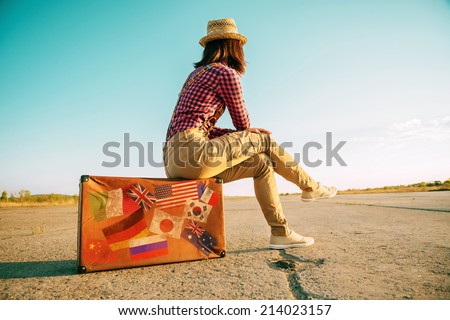 Traveler woman sits on retro suitcase and looks away on road. Suitcase with stamps flags representing each country traveled. Royalty-Free Stock Photo #214023157