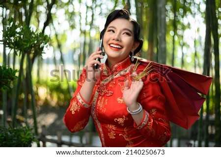Portrait pretty Asian woman in a Chinese cheongsam holding shopping bags and talking on mobile phone with happiness in bamboo forest background, Chinese New Year concept