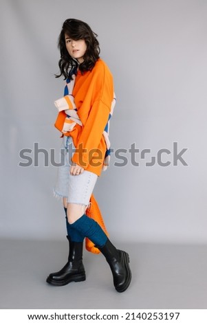 stylish woman dressed in orange knitted jacket posing for a photo in a photo studio on a gray paper background. Bright emotions, poses, modern concept Royalty-Free Stock Photo #2140253197