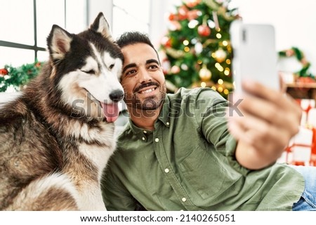 Young hispanic man make selfie by the smartphone sitting on sofa with dog by christmas tree at home