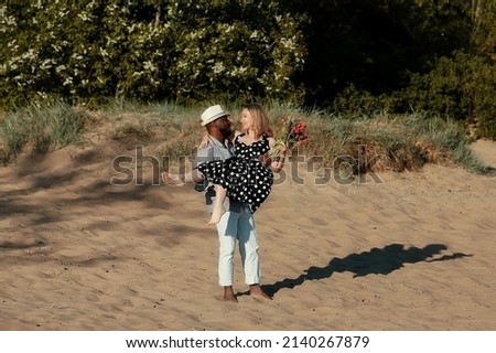 The African American guy carries his girlfriend in his arms. The girl has a large bouquet of tulips in her hands. Romantic date on a summer day at the beach Royalty-Free Stock Photo #2140267879