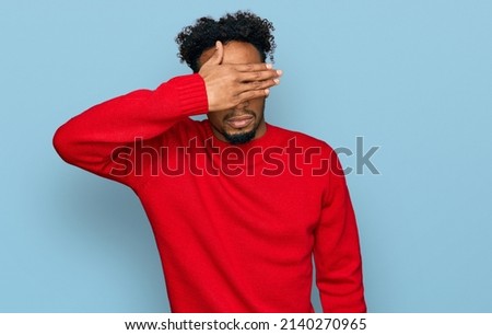 Young african american man with beard wearing casual winter sweater covering eyes with hand, looking serious and sad. sightless, hiding and rejection concept 