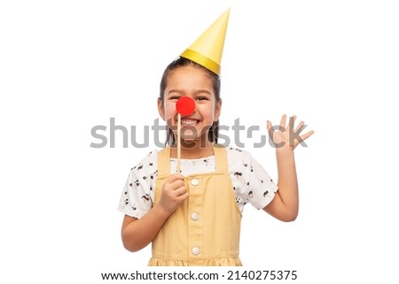 birthday, childhood and people concept - portrait of little girl in dress and party hat with red clown nose over white background