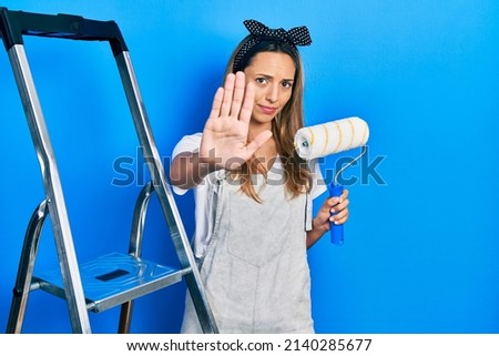 Beautiful hispanic woman by stairs holding roller painter with open hand doing stop sign with serious and confident expression, defense gesture 
