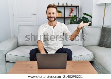 Middle age man using laptop at home smiling cheerful presenting and pointing with palm of hand looking at the camera. 