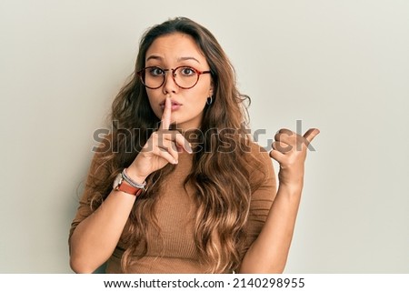 Young hispanic girl wearing casual clothes and glasses asking to be quiet with finger on lips pointing with hand to the side. silence and secret concept. 
