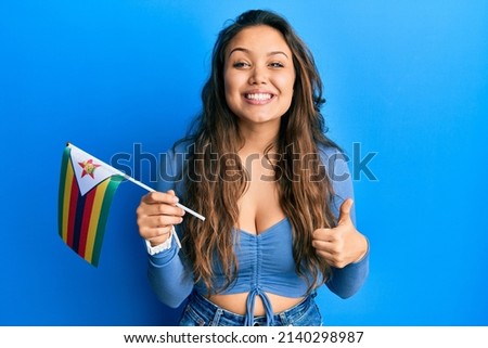Young hispanic girl holding zimbabwe flag smiling happy and positive, thumb up doing excellent and approval sign 