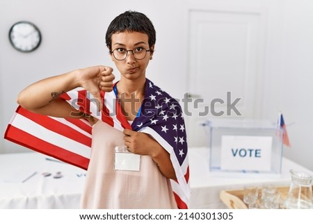 Young hispanic woman with short hair at political campaign election holding usa flag with angry face, negative sign showing dislike with thumbs down, rejection concept 