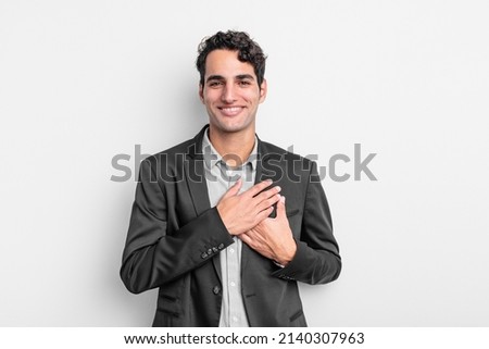 young businessman feeling romantic, happy and in love, smiling cheerfully and holding hands close to heart