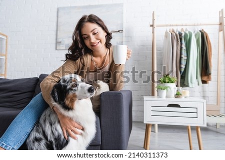 joyful young woman cuddling australian shepherd dog and holding cup of coffee Royalty-Free Stock Photo #2140311373