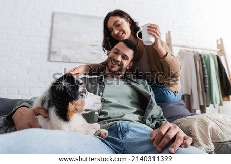 smiling young woman holding cup near bearded boyfriend hugging australian shepherd dog Royalty-Free Stock Photo #2140311399