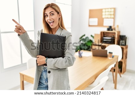 Blonde business woman at the office with a big smile on face, pointing with hand finger to the side looking at the camera. 