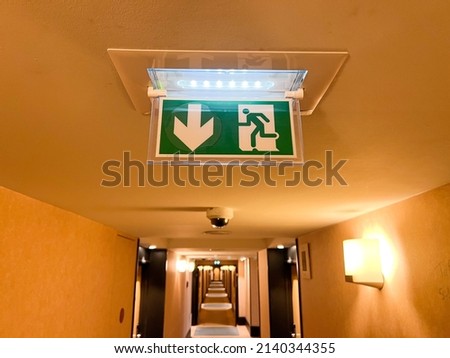 Emergency exit sign at the corridor in building. Green fire exit sign hanging on ceiling on dark corridor in building near fire emergency exit door. Green emergency exit sign. 