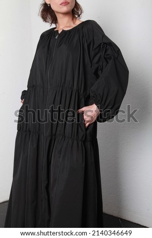 Cropped front side image of unrecognizable female wearing oversize loose black dress coat. Studio shoot of catalog clothes at white walls background. Latest streetwear style trends of spring summer   Royalty-Free Stock Photo #2140346649
