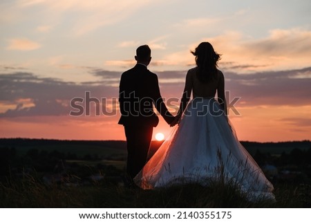 Silhouette of a wedding couple at sunset. Bride and groom in field with sunlight. Portrait of newlyweds enjoying romantic moments. Man and woman holding hands on a meadow in mountains top. Back view.