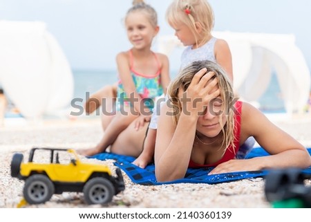 Two cute little sibling girls enjoy having fun playing sitting on tired exhausted mother's back at sea ocean beach. Frustrated mom make face palm gesture. Vacation family small kids trouble concept Royalty-Free Stock Photo #2140360139