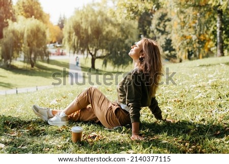 Young caucasian woman resting carefree, relaxing on lawn on sunny day. Side view hipster woman with closed eyes enjoying calmness and freedom during coffee break while sitting on grass outdoors. Royalty-Free Stock Photo #2140377115