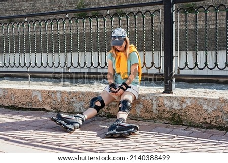 Young smiling woman in cap dressed in roller skates and protective equipment using smartphone chatting while sitting on the embankment