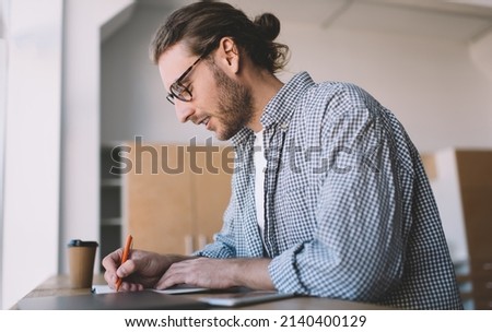 Side view of intelligent male student in optical eyewear writing education information while doing homework task, smart hipster guy in spectacles making notes while preparing to university exams