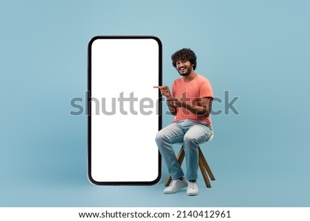Positive curly young indian guy in casual sitting on chair by huge cell phone, pointing at white empty screen and smiling, blue studio background, mockup, panorama with copy space