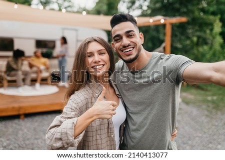 Loving young multiracial couple taking selfie near motorhome, resting with their friends outdoors. Arab guy and his Caucasian girlfriend enjoying summer camping trip, making mobile photo