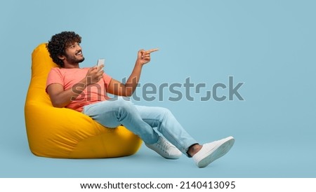 Happy curly millennial indian man sitting at bean bag with brand new smartphone and pointing at copy space over blue studio background, showing nice offer and smiling, panorama Royalty-Free Stock Photo #2140413095
