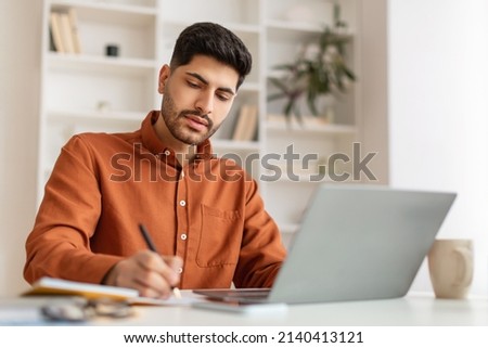 Focused young Middle Eastern man sitting at desk working on laptop taking notes in notebook, busy millennial male studying online, watching webinar using computer and writing check list Royalty-Free Stock Photo #2140413121