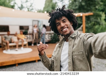 Happy black guy taking selfie near RV, smiling at camera, resting with his multiracial friends outdoors. Cool African American teenager enjoying camping trip, making mobile photo, copy space