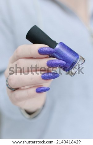 Female hand with long nails and purple plum blue manicure holds a bottle of nail polish