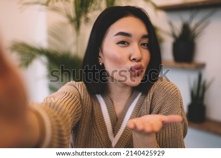 Portrait of asian girl with air kiss gesture taking selfie in blurred cafe. Partial image of young beautiful millennial brunette woman. Concept of rest, leisure and free time. Modern female lifestyle